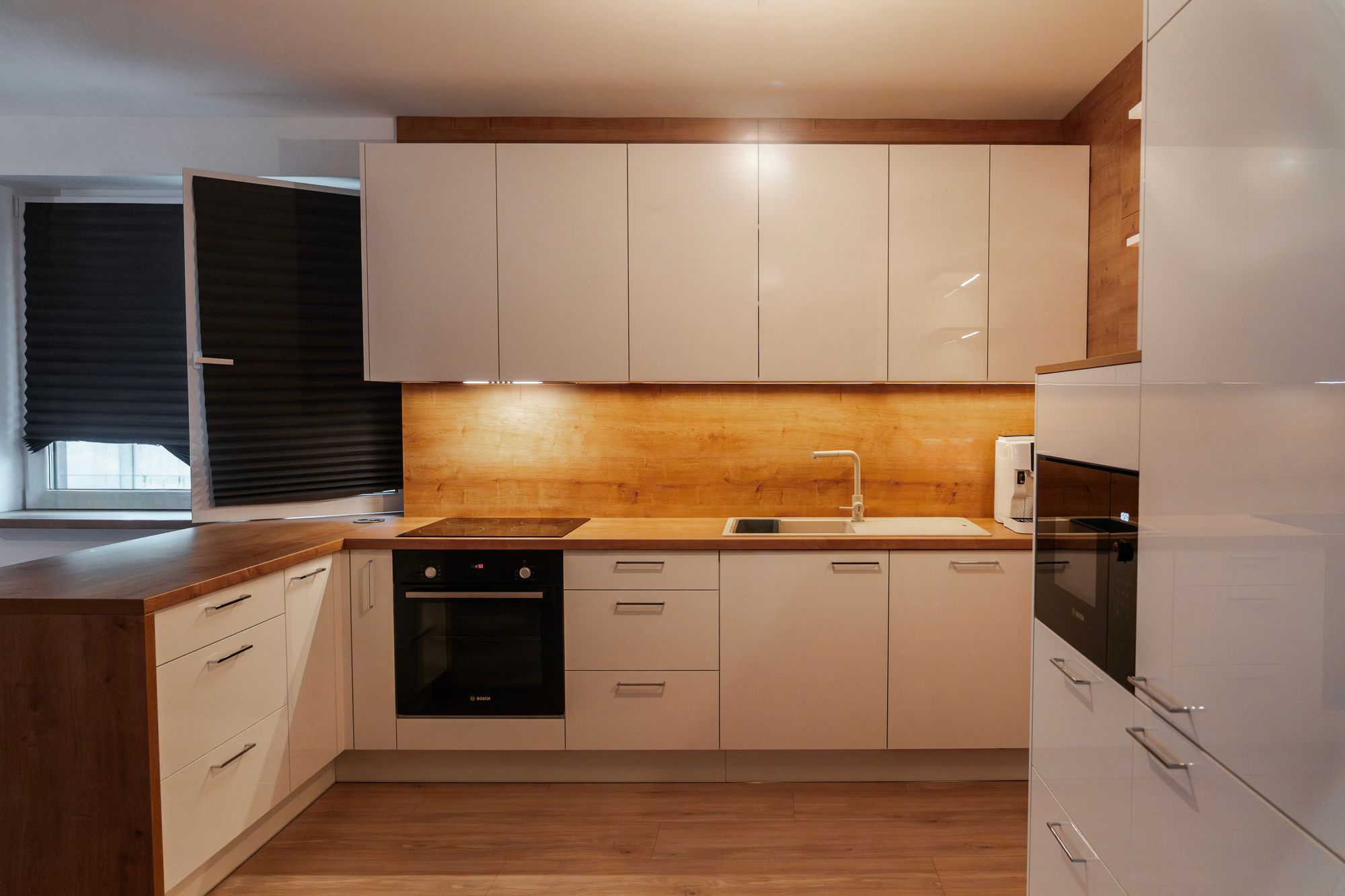 U-shaped kitchen with gloss white cabinets, worktop and splashback in Hamilton oak decor, built-in oven and hob, Veľký Biel.