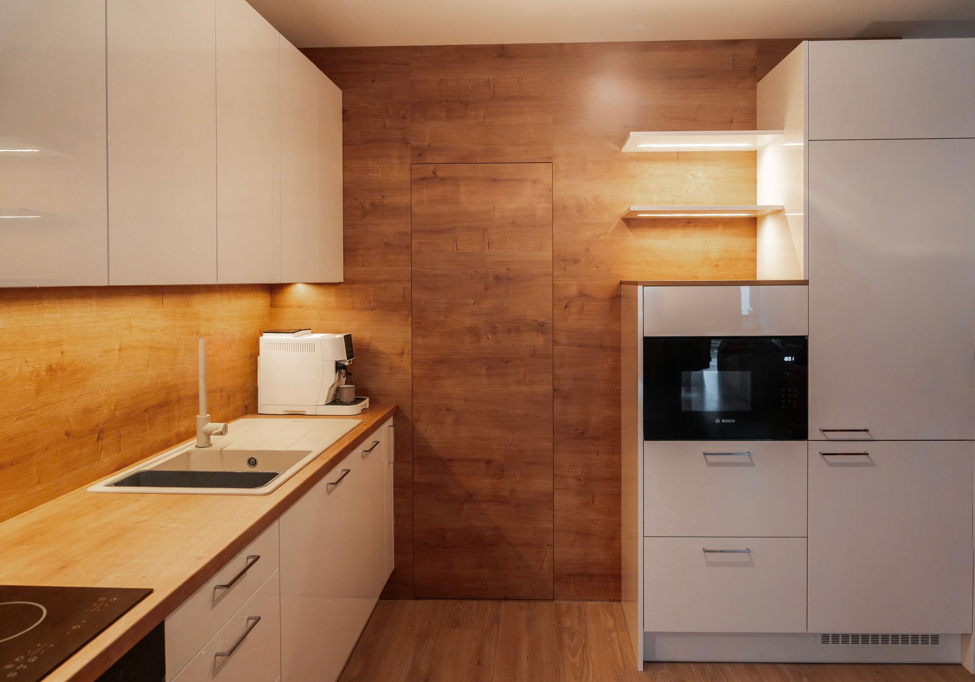 Gloss white kitchen with a built-in oven and microwave, natural Hamilton oak panelling and open illuminated shelves, Veľký Biel.