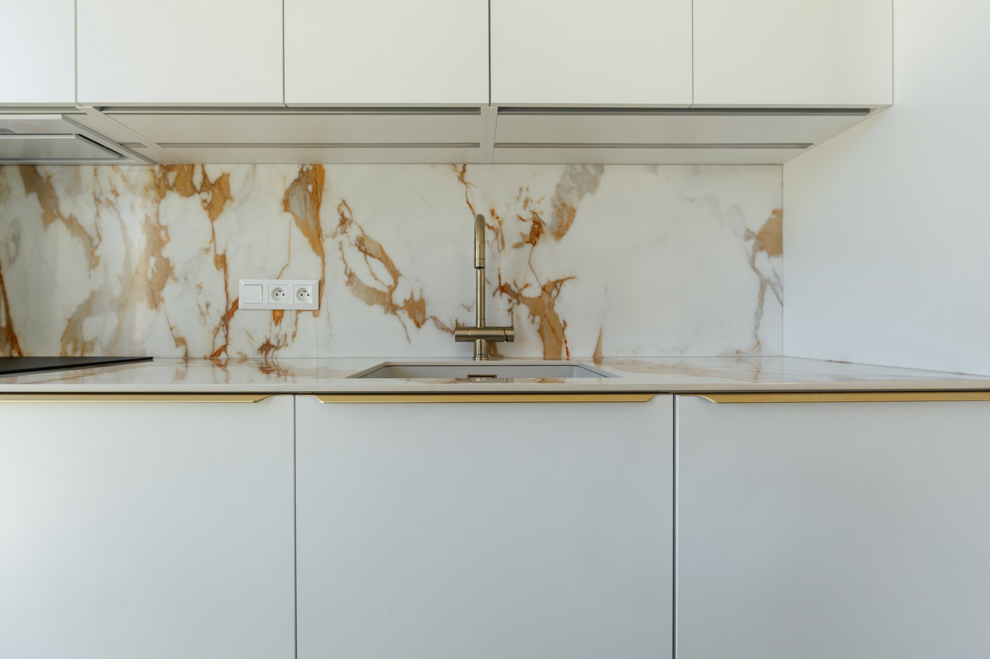 Detail of a white kitchen with a marbled ceramic splashback and worktop, gold tap and handles, Čunovo.